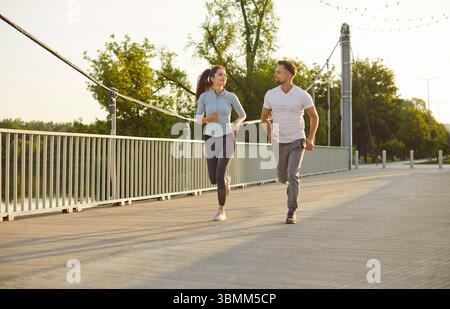 Smiling sporty young couple jogging along city bridge and enjoying outdoor workout together. Stock Photo