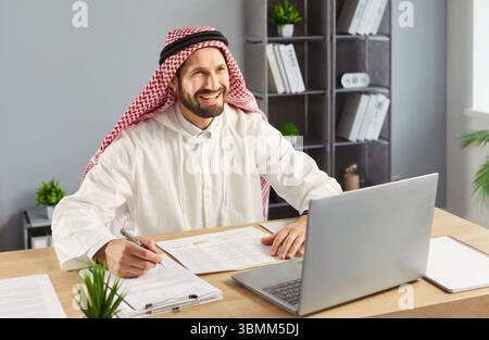 Happy Arabian man working on financial report, writing in paper documents and using laptop Stock Photo