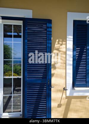 Yellow house facade with open blue shutters on window and balcony door under bright sunlight. Stock Photo