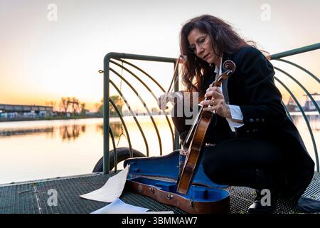 beautiful middle-aged woman dressed in an elegant tailcoat taking a handmade violin out of its case at sunset on a small metal pier Stock Photo
