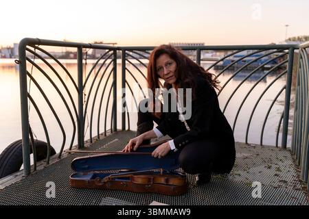 beautiful middle-aged woman dressed in an elegant tailcoat taking a handmade violin out of its case at sunset on a small metal pier Stock Photo