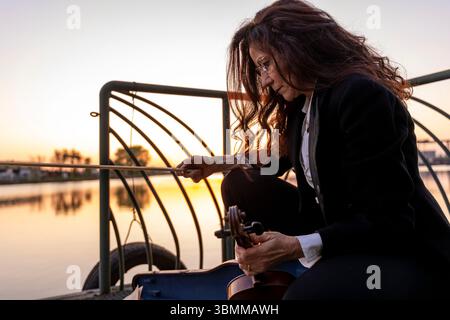 beautiful middle-aged woman dressed in an elegant tailcoat taking a handmade violin out of its case at sunset on a small metal pier Stock Photo
