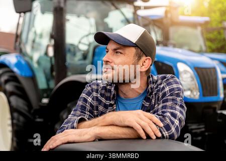 Portrait of smiling male farmer in plaid shirt and cap with blue tractor in background Stock Photo