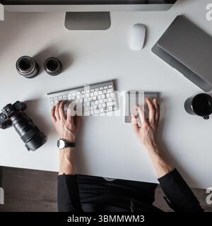 Young Successful Photographer Is Sitting At The Table And Typing On The Keyboard. Top View Of The Desktop On Which There Is A Keyboard, Camera, Laptop Stock Photo
