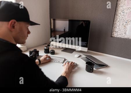 Successful Stylish Young Photographer Is Working Behind A Computer In An Office. Man In The Cap Sits At A Table On Which There Is A Keyboard, Camera, Stock Photo