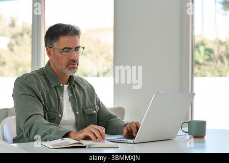 Serious mature older man using laptop computer at home table in living room. Stock Photo