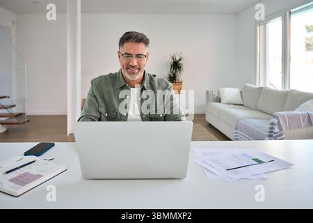 Happy older middle aged man using laptop computer at home table. Stock Photo