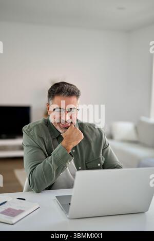 Happy middle aged man using laptop computer writing notes sitting at home table. Stock Photo
