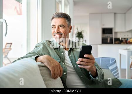 Relaxed older man using phone sitting at home on sofa looking away. Stock Photo