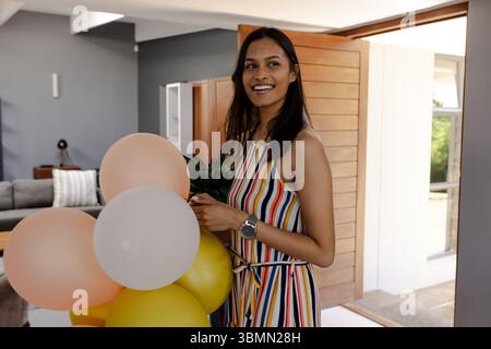 Smiling woman holding peach white balloon cluster by open wooden door at home wearing striped dress Stock Photo