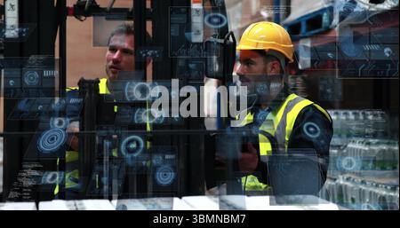 Two men wearing vests, helmet operating forklift controls in warehouse with shelving and pallets Stock Photo