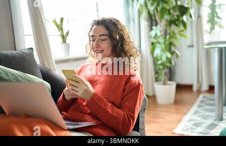 Smiling young woman sitting on couch holding phone in cozy living room interior. Stock Photo