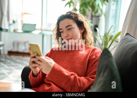 Relaxed young woman sitting on couch using mobile phone technology. Stock Photo