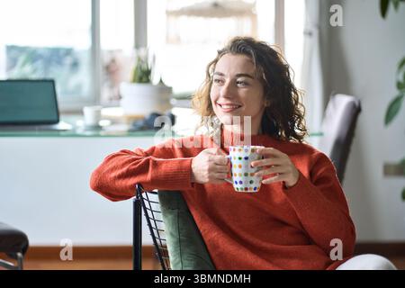 Young happy pretty woman sitting on chair drinking tea coffee at home. Stock Photo