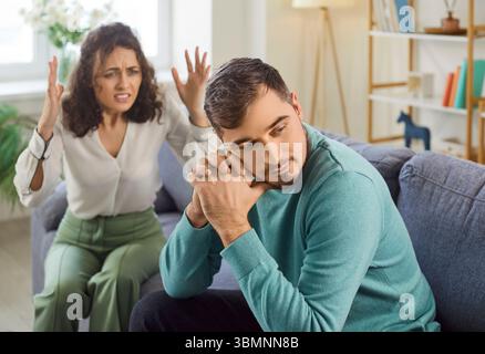 Sad man sitting on sofa while angry woman shouting on him, showing frustration and disappointment. Stock Photo