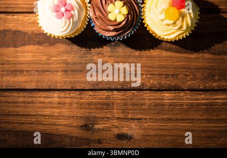 Three frosted cupcakes are resting on wood grain table in flat design with fondant flowers, gummies Stock Photo