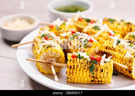 Grilled corn on cob sprinkled with cilantro and grated parmesan in a white dish on a table, close up. Stock Photo