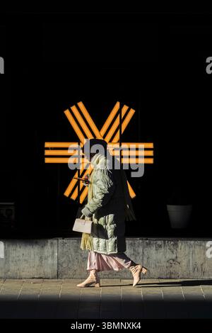 A stylish figure walks past a striking geometric light installation, blending modern fashion with urban aesthetics. Stock Photo
