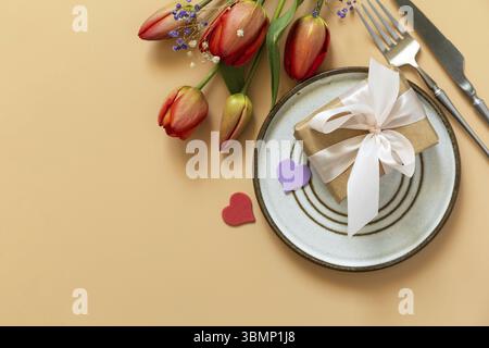 Beautiful table setting with gift box, decorative hearts and tulips flowers on pastel peach background. Happy Mother's day concept background 2024. Vi Stock Photo