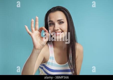 Magnificent brunette lady with a long hair, dressed in a colorful striped shirt is showing ok sign and smiling while posing against a blue studio back Stock Photo