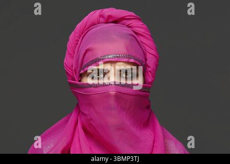 Close-up shot of a young female with a bright make-up wearing the pink hijab decorated with sequins. She is posing at the studio and looking angry on Stock Photo