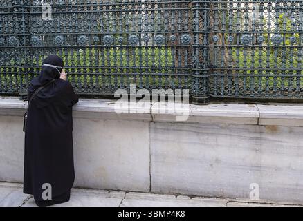 Muslim woman in black dress and headscarf is praying looking through a metal fence outside a mosque in Istanbul Stock Photo