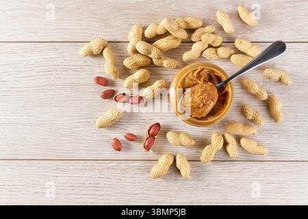 peanut paste in an open jar and peanuts in the peel scattered on wooden table. Stock Photo
