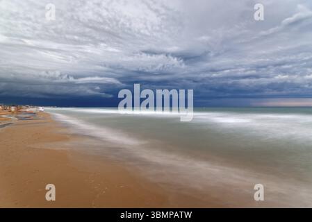 A long exposure captures the motion of the waves as they meet the shore under a dramatic, cloudy sky at the beach. Cut-off low Stock Photo