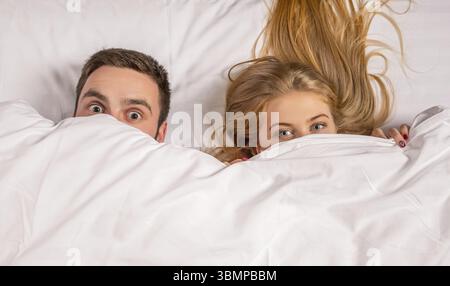 Young lovely couple lying and have fun in a bed, happy smile looking at camera, cover under blanket. Family, bedtime and happiness concept Stock Photo