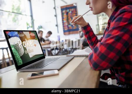 Side view of a young pink hair woman keyboarding on laptop computer with poker online on a screen while sitting in cafe. Play online concept. Screen g Stock Photo