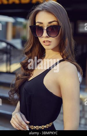 Fashion style portrait of young beautiful elegant woman in black dress, sunglasses, with long dark hair walking at city streets Stock Photo