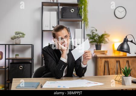 Serious Caucasian young businessman in formal suit sitting at home office with letter, upset and disappointed, reading bad news, receiving notification of bank loan refusal, frustrated by bankruptcy. Stock Photo