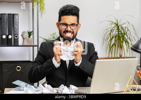 Angry furious Indian man working at office throwing crumpled paper, having nervous breakdown at work, migraine headache, stress management, mental distress problems, losing temper, reaction on failure Stock Photo