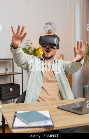Confident Caucasian man freelancer testing VR headset while sitting at home office table. Young businessman guy making presentation using new gadget during remote working online at home. Vertical. Stock Photo