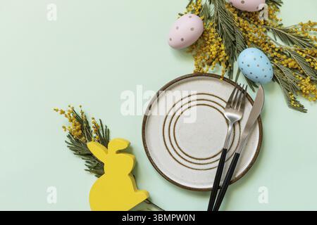 Easter table setting. Plate with easter eggs and mimosa on a pastel green background. Happy Easter holiday concept for restaurants. View from above. C Stock Photo
