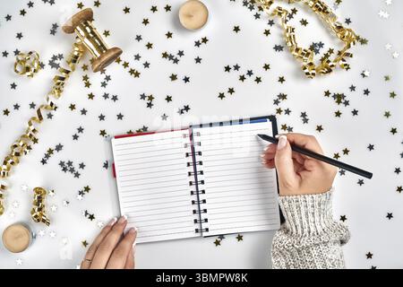 Festive decorations, delicate wavy ribbons, metallic star shaped confetti and notebook with wish list on white table, flat lay style. Planning concept Stock Photo