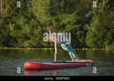 Photo of young woman doing hand stand on stand up paddle board. She wearing a leggings and top. Sunny day, blue lake and green trees on the shore Stock Photo