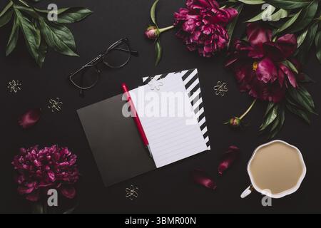 Female Home office desk. Elegant Workspace with notebook, accessories, coffee cup and peony flowers on black background Stock Photo