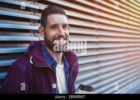 Handsome young man in purple winter jacket drinking coffee. The guy drinking coffee on the street. Man holding paper cup with americano or latte. Stre Stock Photo