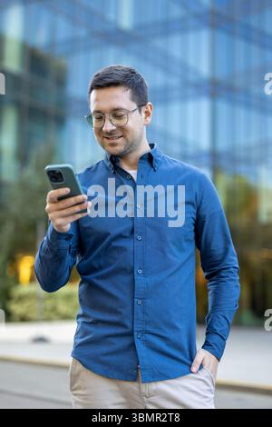 A man in glasses is using a smartphone outside in front of a modern office building, he seems pleased. Stock Photo