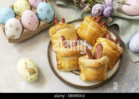 Buns in the form of an Easter rabbit from sausage and cheese in a yeast dough with colored eggs on a gray stone background. Traditional Easter symbols Stock Photo