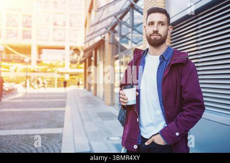 Handsome young man in purple winter jacket drinking coffee. The guy drinking coffee on the street. Man holding paper cup with americano or latte. Stre Stock Photo