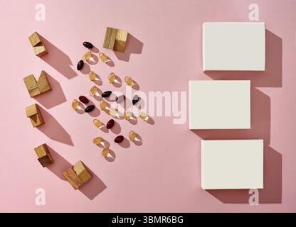 Three small white cardboard boxes with no logo, scattered colorful capsules and wooden blocks of different shapes on pink studio background. Close up Stock Photo