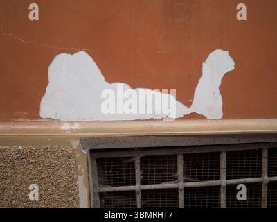 Weathering paint exposing white plaster over basement window with rusty metal grate, historic building facade in Cremona, Lombardy Stock Photo