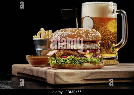 Hamburger with two juicy patties, fried bacon, cheese, caramelized onions, fresh tomatoes and lettuce in fluffy sesame bun served on wooden board with Stock Photo