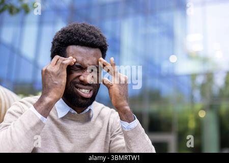 A man experiencing a headache, pressing his fingers to his temples with eyes closed outdoors. Stock Photo