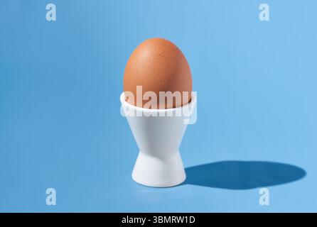 One brown chicken egg stands in a white egg-cup on a blue background. Hard light, copy space, close-up. Stock Photo