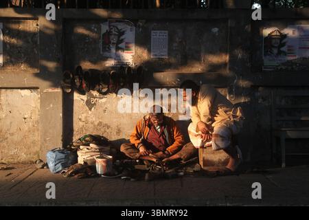 Dhaka Bangladesh 27 December 2024, Life's struggle between light and shadow , A cobbler is repairing shoes on the sidewalk, with a customer sitting ne Stock Photo