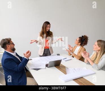 Cheerful group of engineers with blueprints and laptop on table applauding for successful female leader in meeting Stock Photo