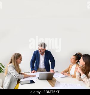 Smiling male project manager with blueprints and computer on table sharing ideas with coworkers while collaborating in meeting Stock Photo